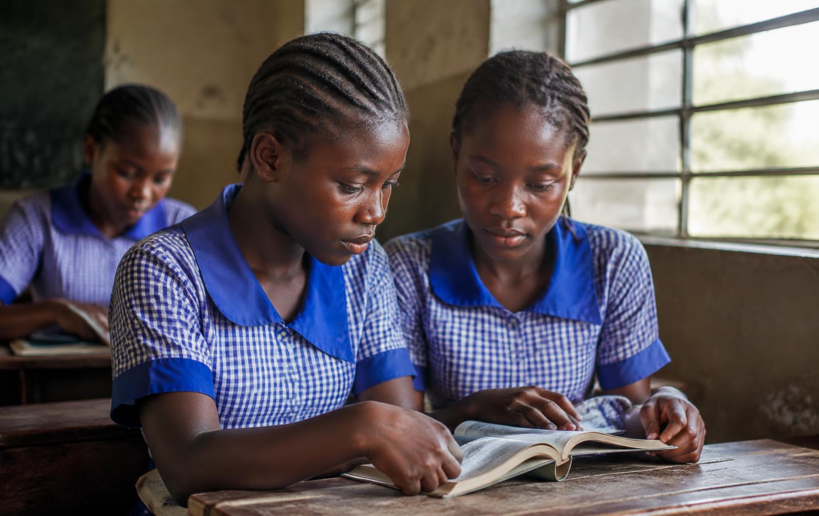 Two schoolgirls reading a book together at a classroom desk